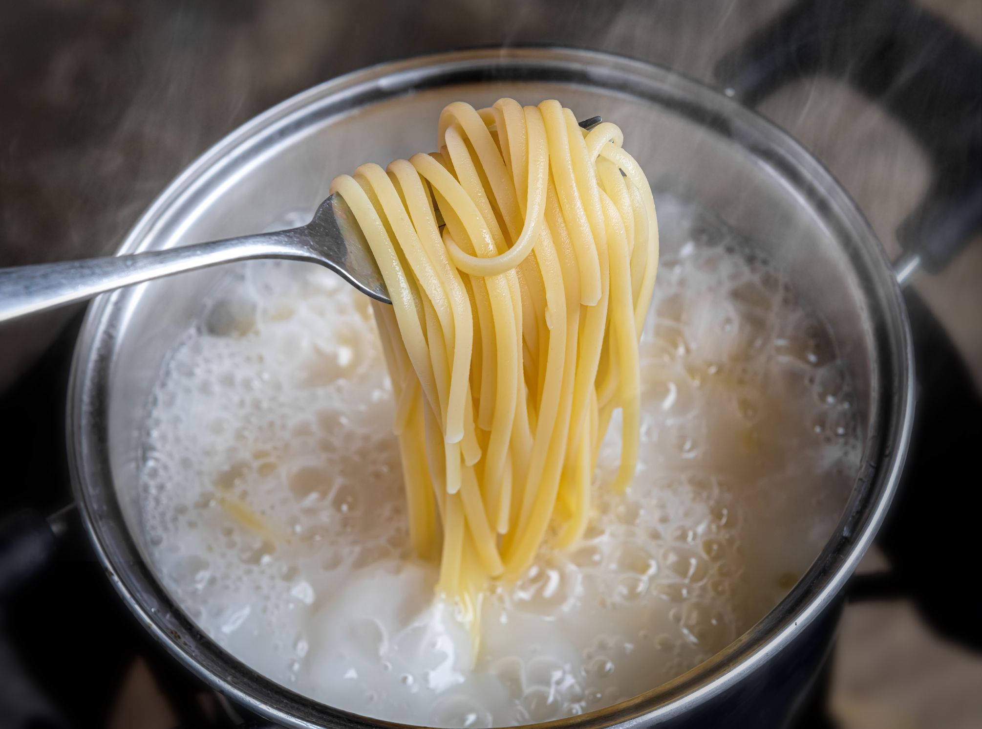 Yellow noodles or spaghetti cooking in boiling water pot with indoor low lighting.