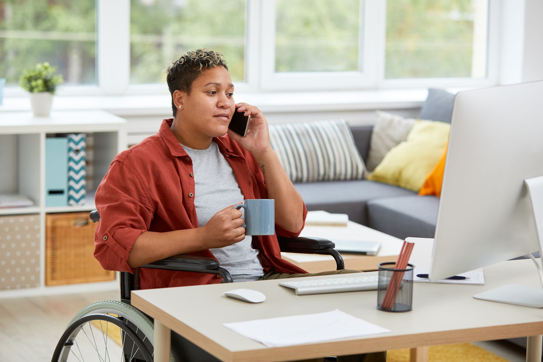 woman sitting in wheelchair at the table and looking at computer monitor while talking on mobile phone and drinking coffee at home