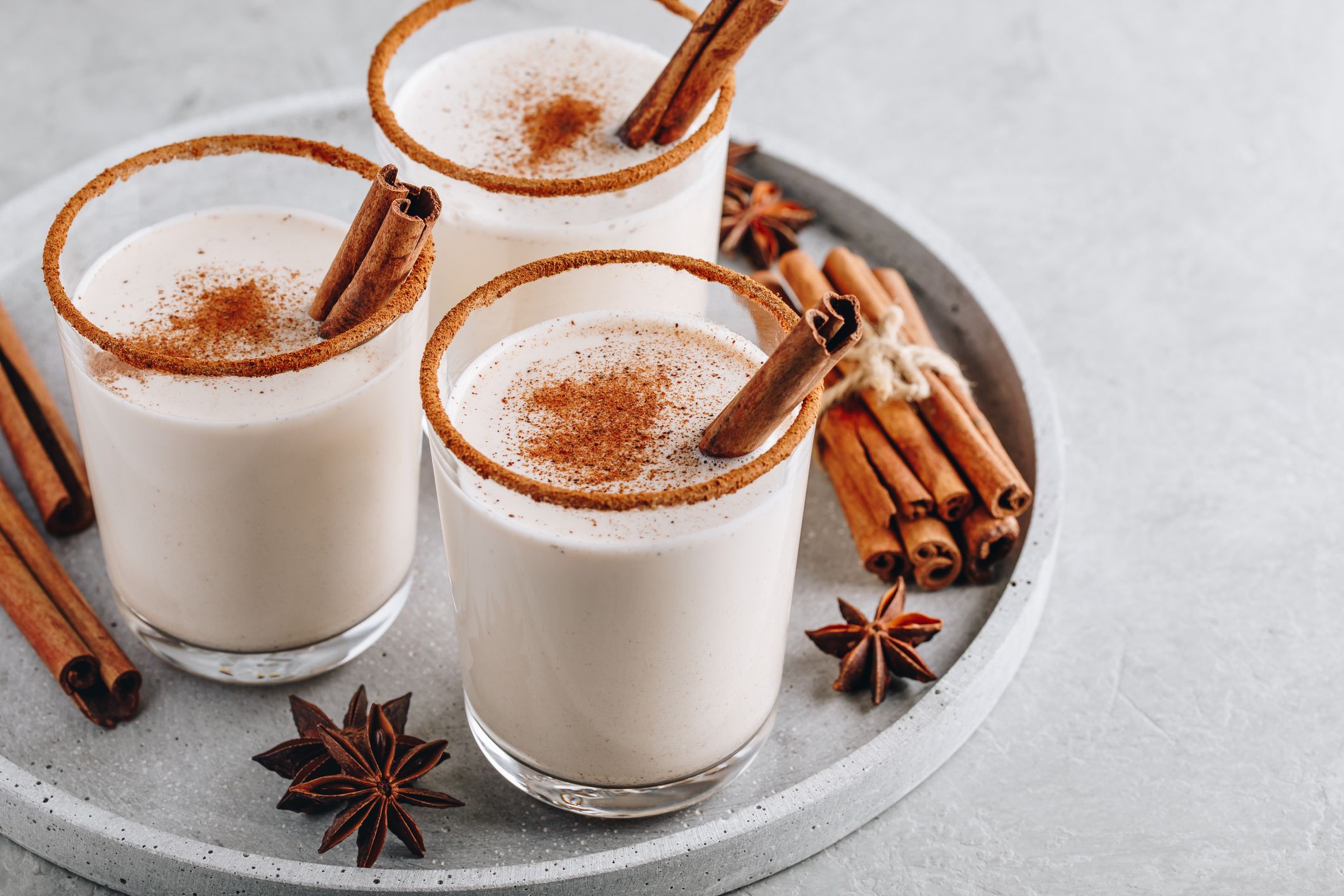 Homemade vanilla Christmas drink Eggnog in glass with grated nutmeg and cinnamon sticks on gray stone background.