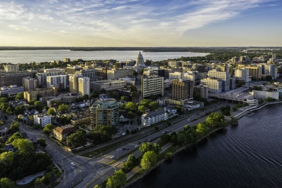 Aerial view of a city with a domed capitol building in the center, surrounded by various buildings, roads, trees, and a large body of water under a partly cloudy sky.