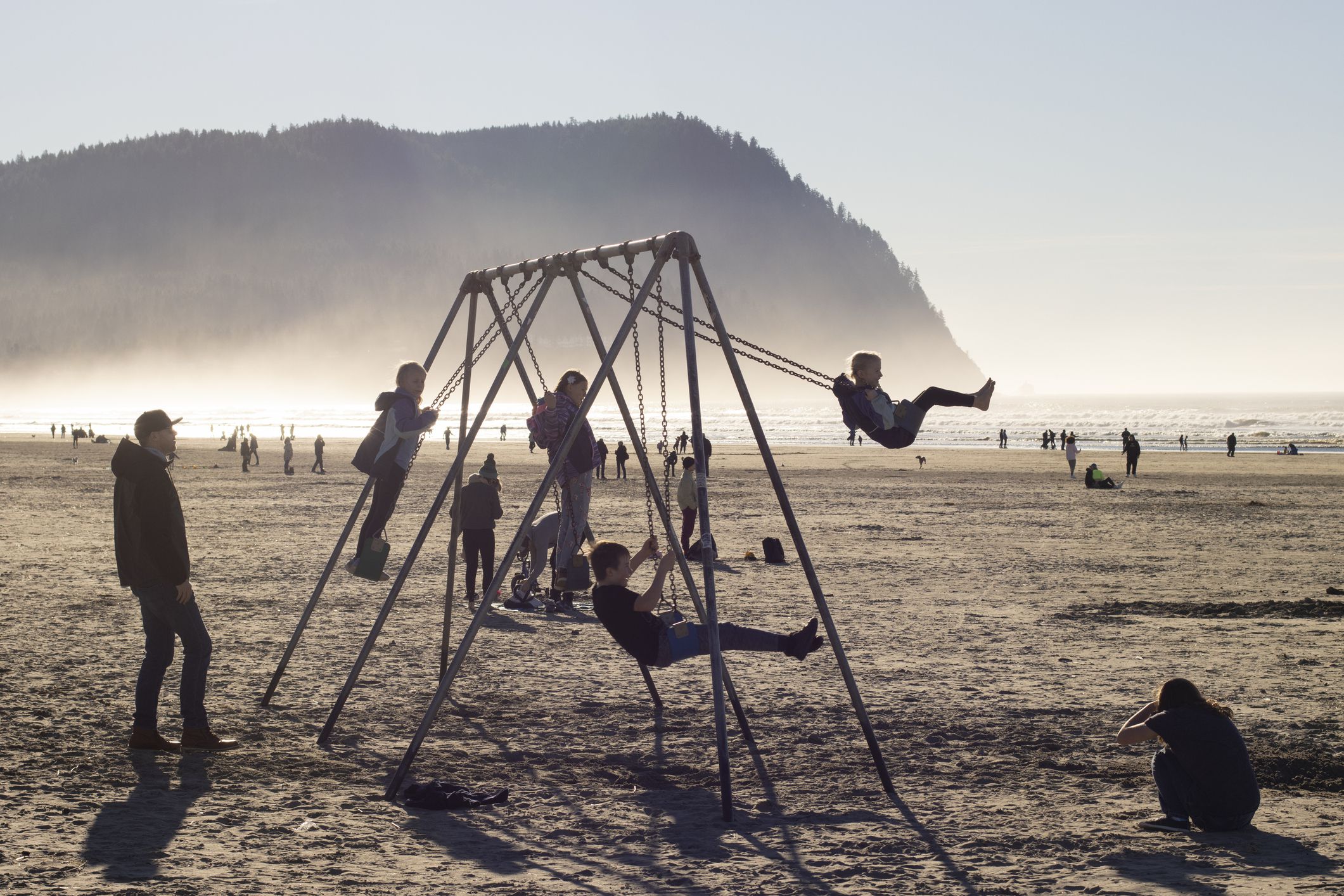 Seaside, OR, USA - Jan 22, 2022: Children play on the beach swings facing the Pacific Ocean in Seaside, Oregon, with parents watching by the side, on a warm winter day.