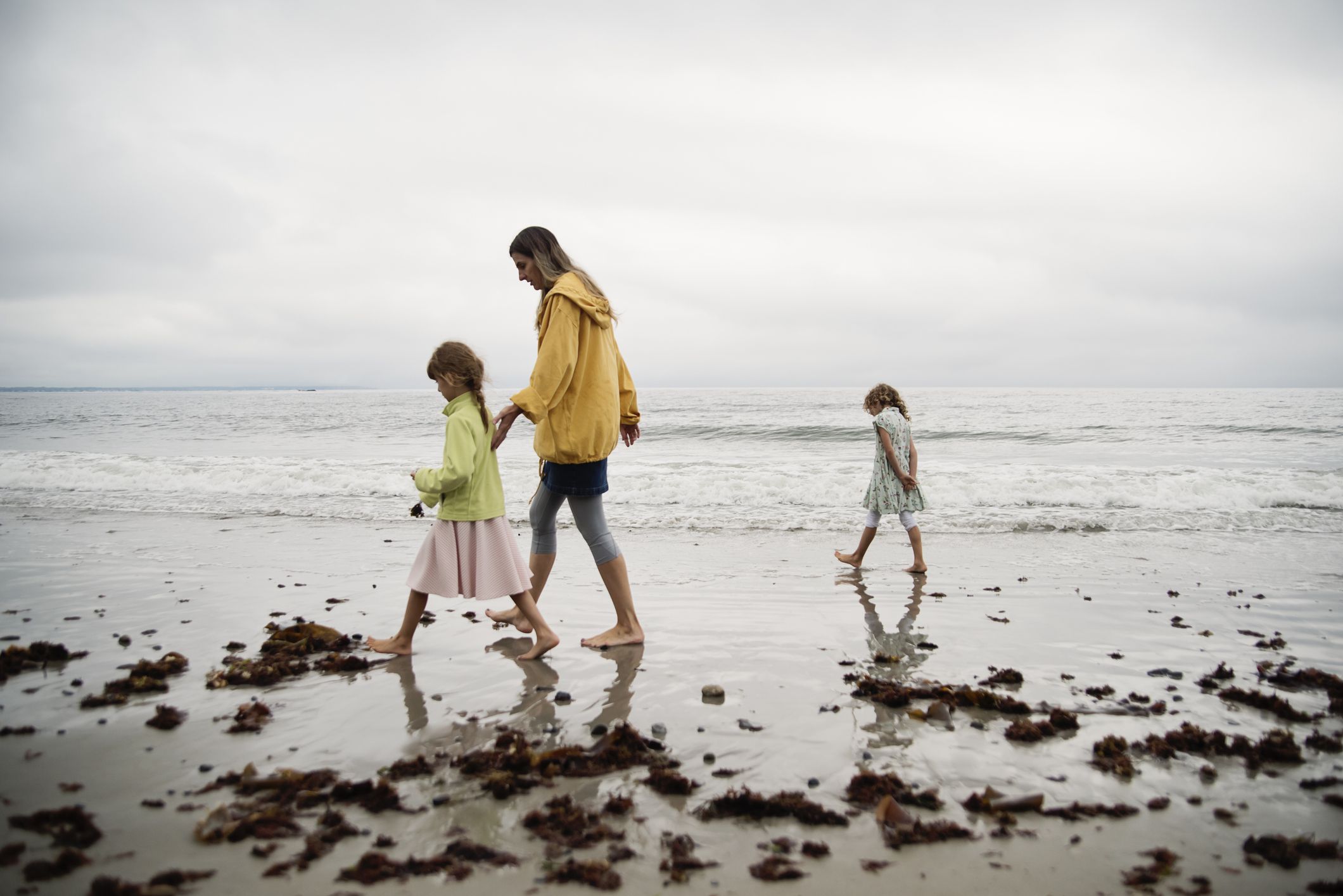 Mother and daughters walking on the beach on an overcast day. End of summer chilly day, off-season holidays. Mom is in her forties, girls are 6 and 8 year’s old. Horizontal outdoors full length shot with copy space.
