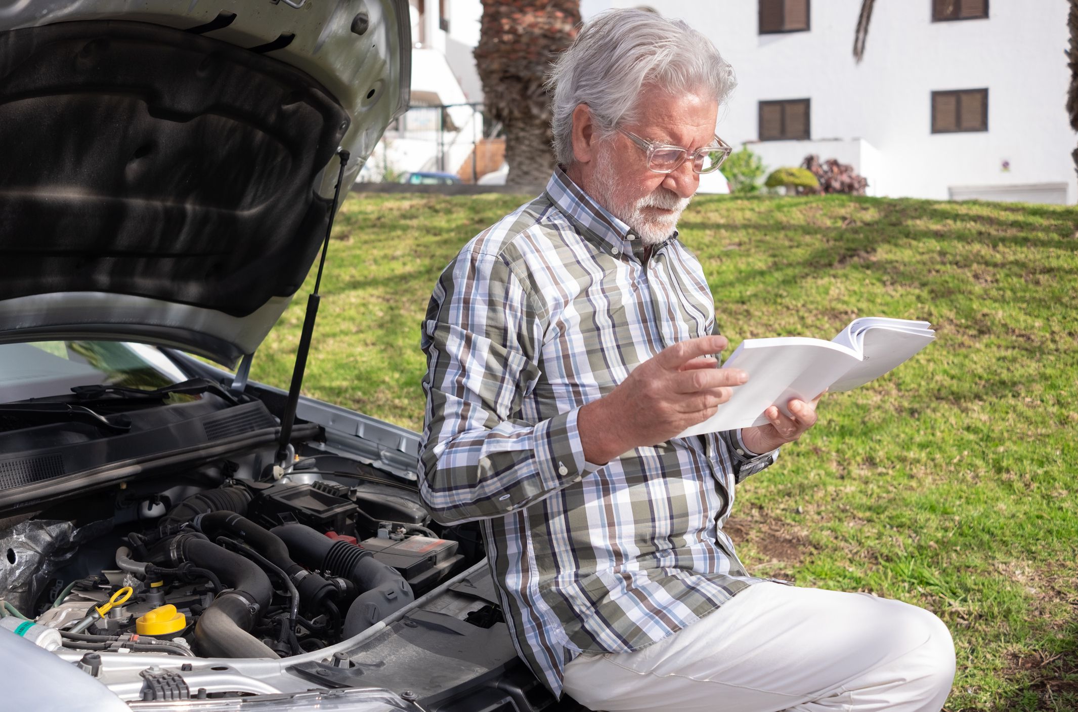 Senior man driver stopped in the street for a car breakdown, the hood open waiting for roadside assistance. Looking at the instruction booklet