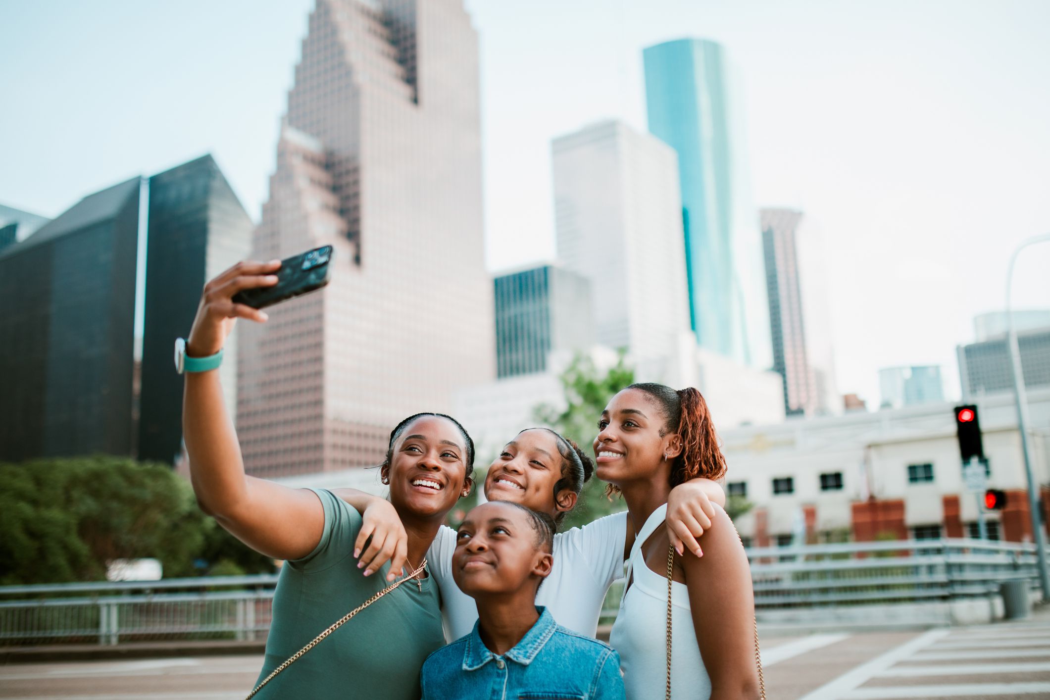 A Black mother and her daughters have fun traveling around the city of Houston, Texas. They walk around the city, skyscrapers visible in the background, pausing to take pictures of the surroundings and themselves.