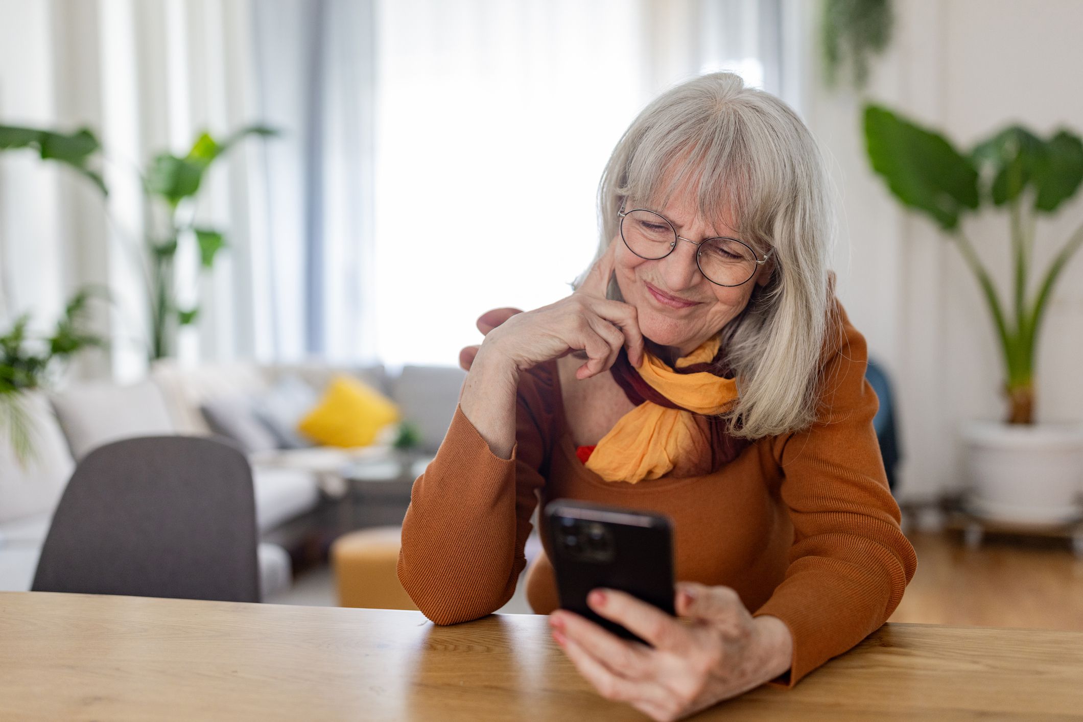 Senior woman skillfully navigates her smartphone at the table, embracing the benefits of digital connectivity and entertainment in her daily life.