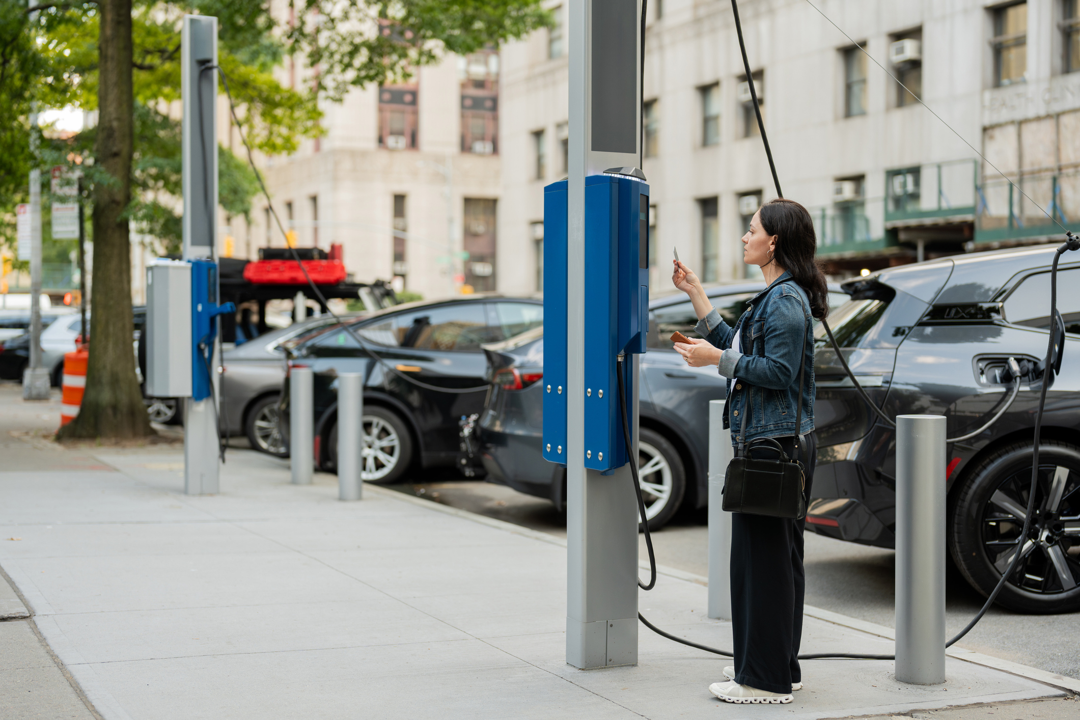 A woman stands on a city sidewalk using an electric vehicle charging station, with several parked cars charging nearby and buildings visible in the background.