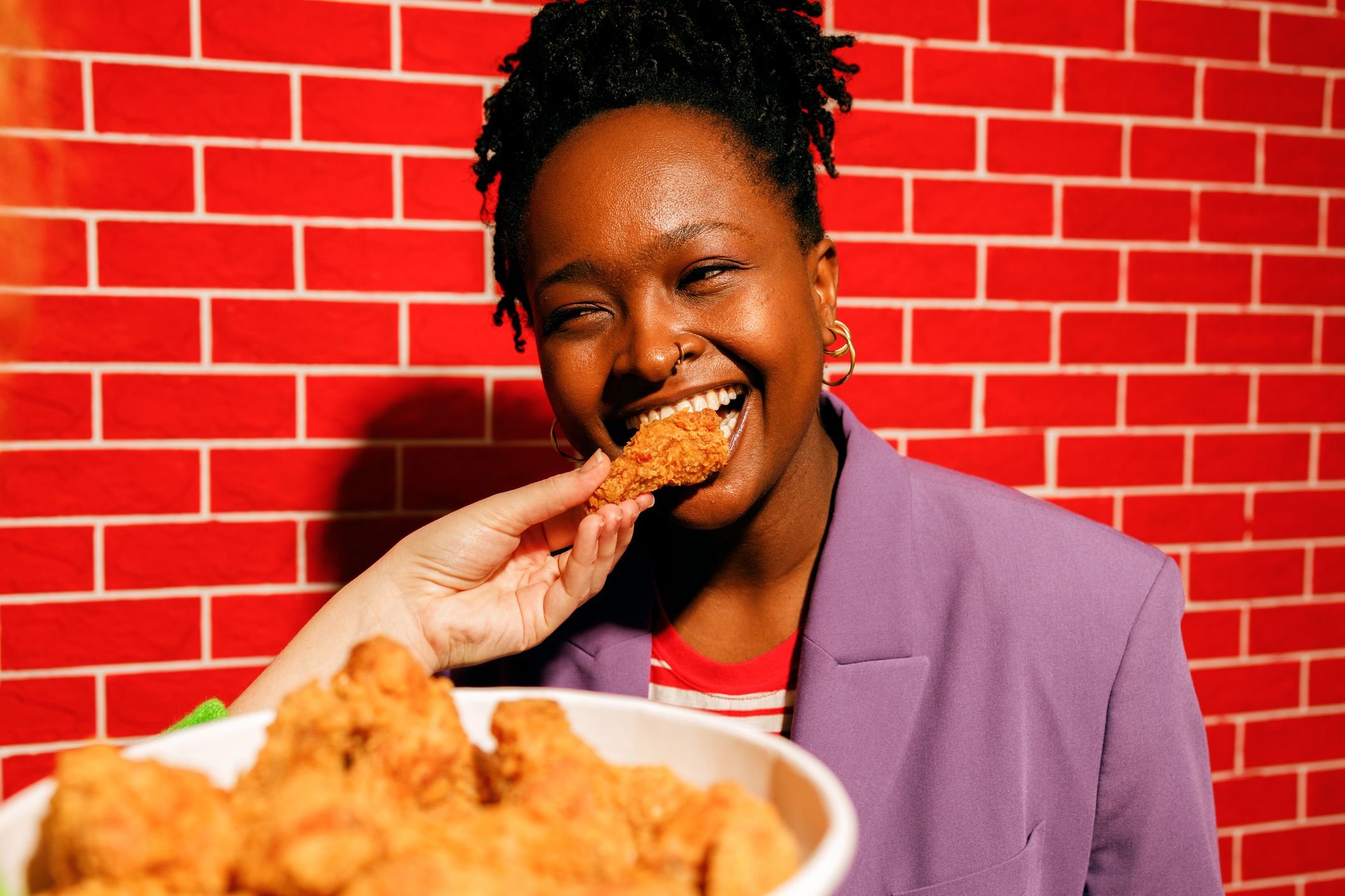 A joyful young black woman in a stylish purple blazer laughs as her friend playfully feeds her a crispy fried chicken piece. A large bowl of golden, crunchy fried chicken is in the foreground, emphasizing the delicious fast food theme.