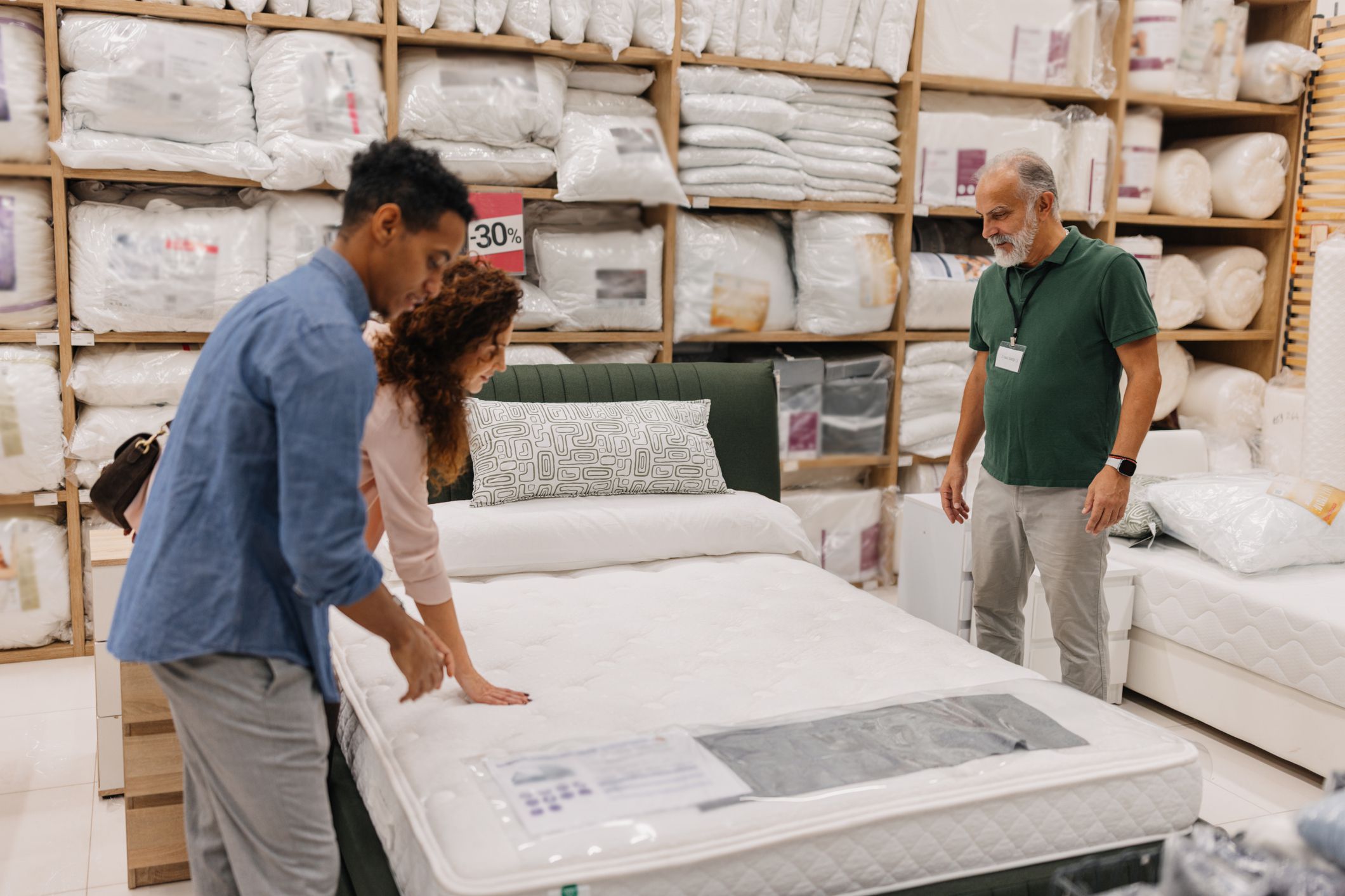 A couple is testing a mattress, with one participant feeling its surface, emphasizing the tactile experience and shared decision-making in their shopping journey.