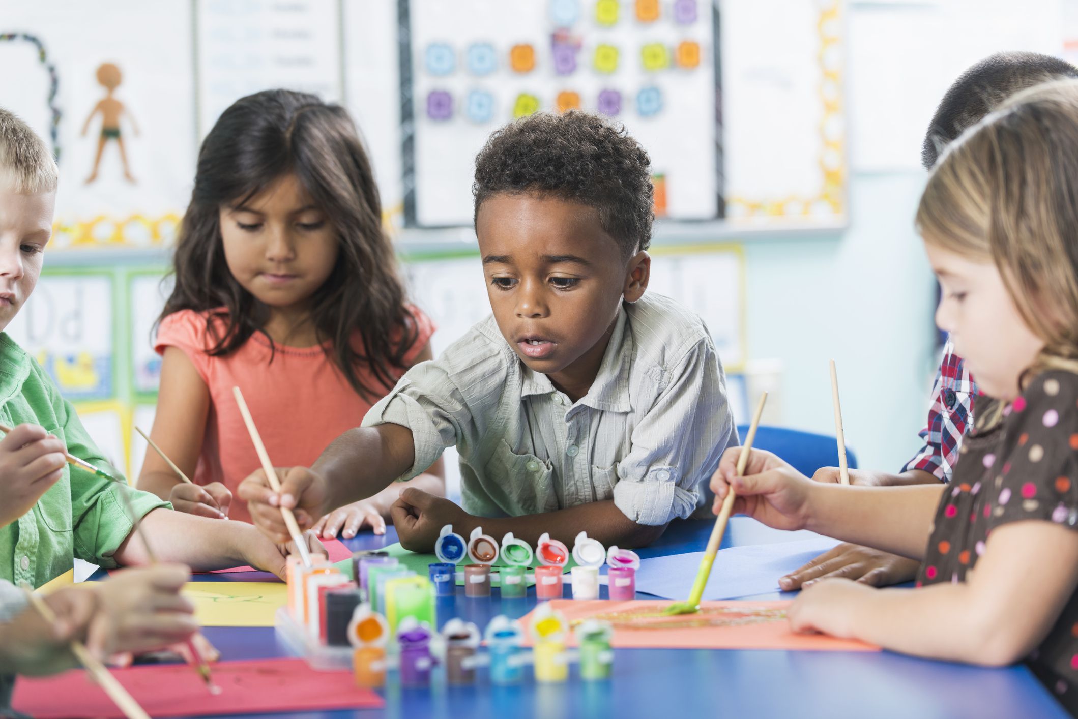 A group of preschool or kindergarten children in art class painting pictures at a table. A little African American boy, 6 years old, is in the middle, reaching over to dip his brush in the paint.