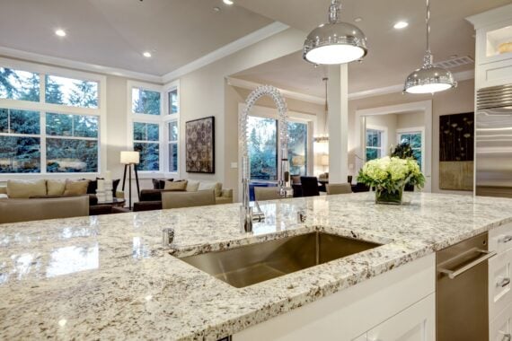 Modern kitchen with granite island, stainless steel sink, pendant lights, and a flower vase. Open layout connects to a bright living area with large windows, beige sofas, and contemporary decor.