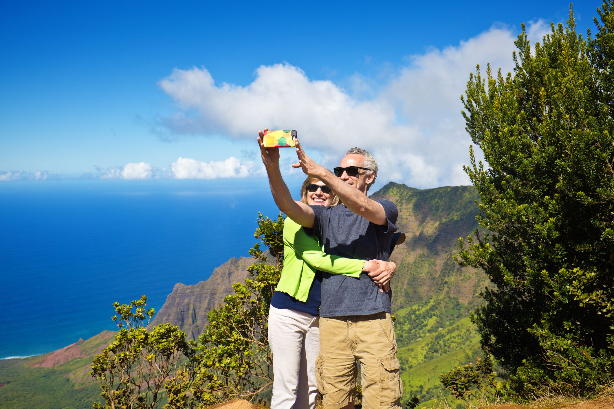 Tourist couple enjoying the scenic view from the Kalalau Lookout in the Waimea Canyon of Kauai, Hawaii. They are holding up smart phone and photographing a selfie with their mobile phone camera.