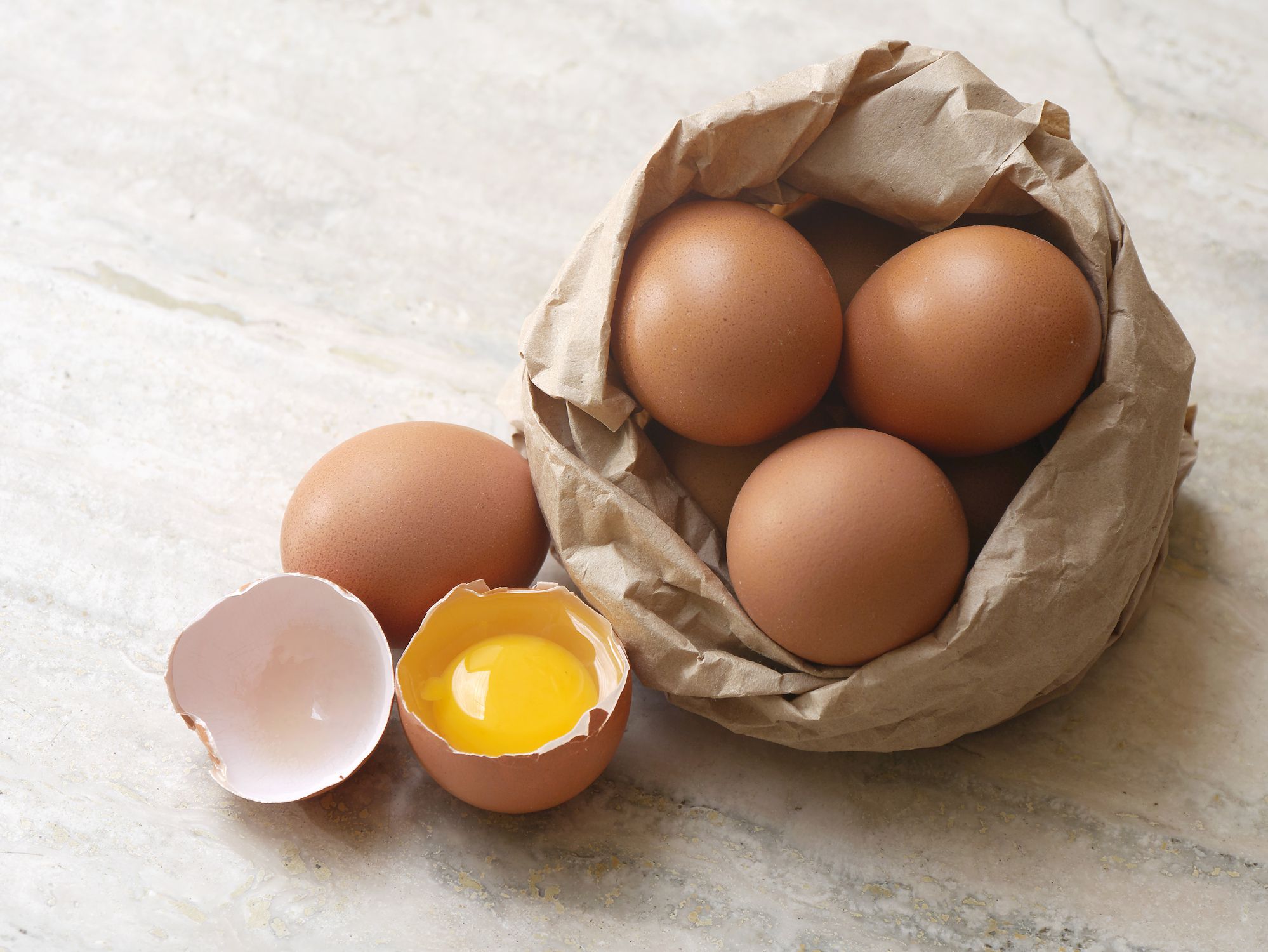 Eggs composition on marble surface, one of them is broken in two halves; some eggs are inside a paperbag. Natural day light horizontal composition with copy space, high angle view. No people. Color photography.