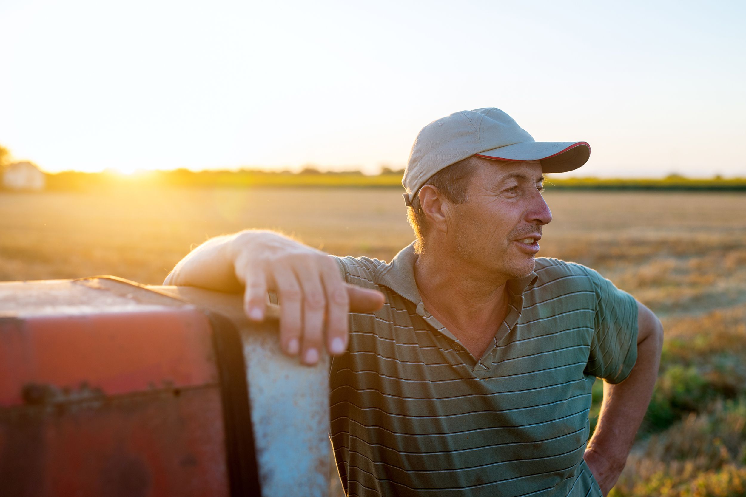 senior agronomist man standing in a field leaning against old tractor after harvest at sunset
