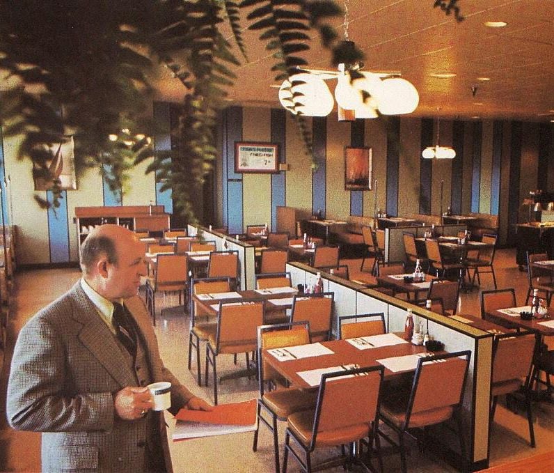 A man in a suit holds a coffee cup, standing in an empty Kmart cafeteria with neatly arranged tables and chairs. The room features striped walls, pendant lights, and hanging plants, creating a retro ambiance.