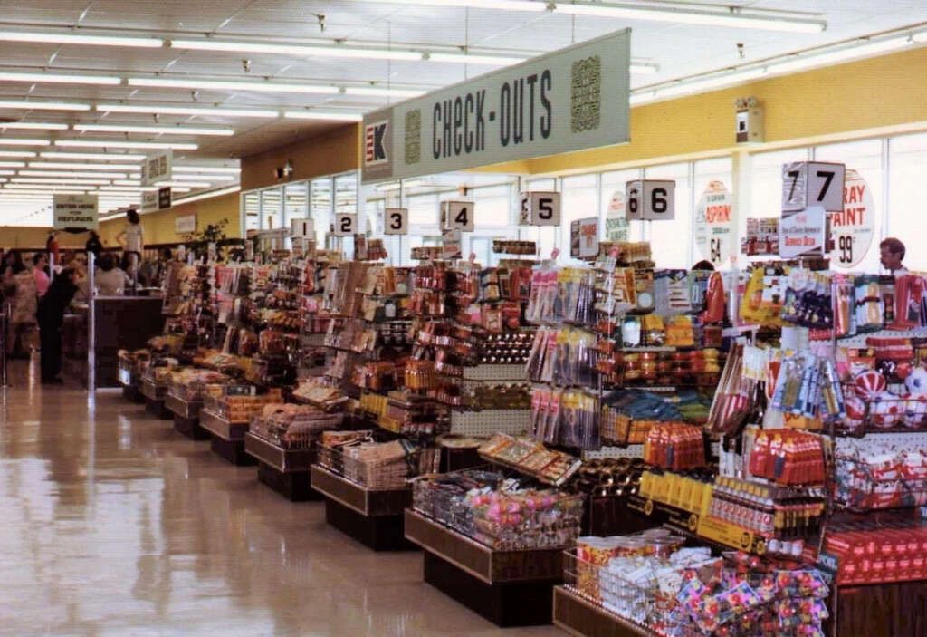 A vintage Kmart checkout area with numbered lanes, stocked with various colorful snacks and candies. Signs hang above the lanes, and shoppers can be seen in the background pushing carts.