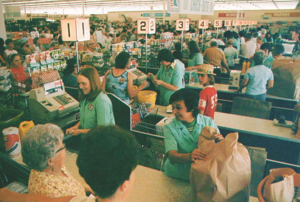 A busy Kmart checkout area with numerous shoppers queuing. Employees in green uniforms assist customers with groceries. A mix of different people and products are visible in a lively atmosphere.