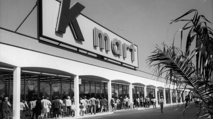 Black-and-white photo of a crowd lined up outside a Kmart store under a large exterior sign, with palm fronds visible on the right side of the image.