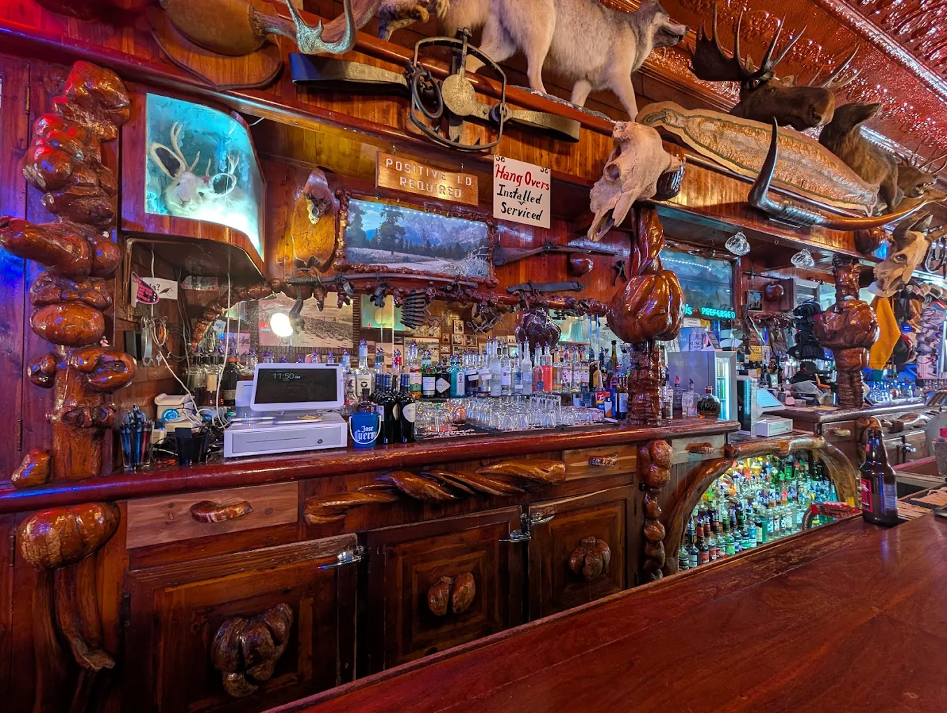 A rustic bar with carved wooden decor, shelves lined with liquor bottles, glasses, and taxidermy animal heads mounted above. A cash register and barware sit on the counter, with a mirror reflecting the cozy interior.