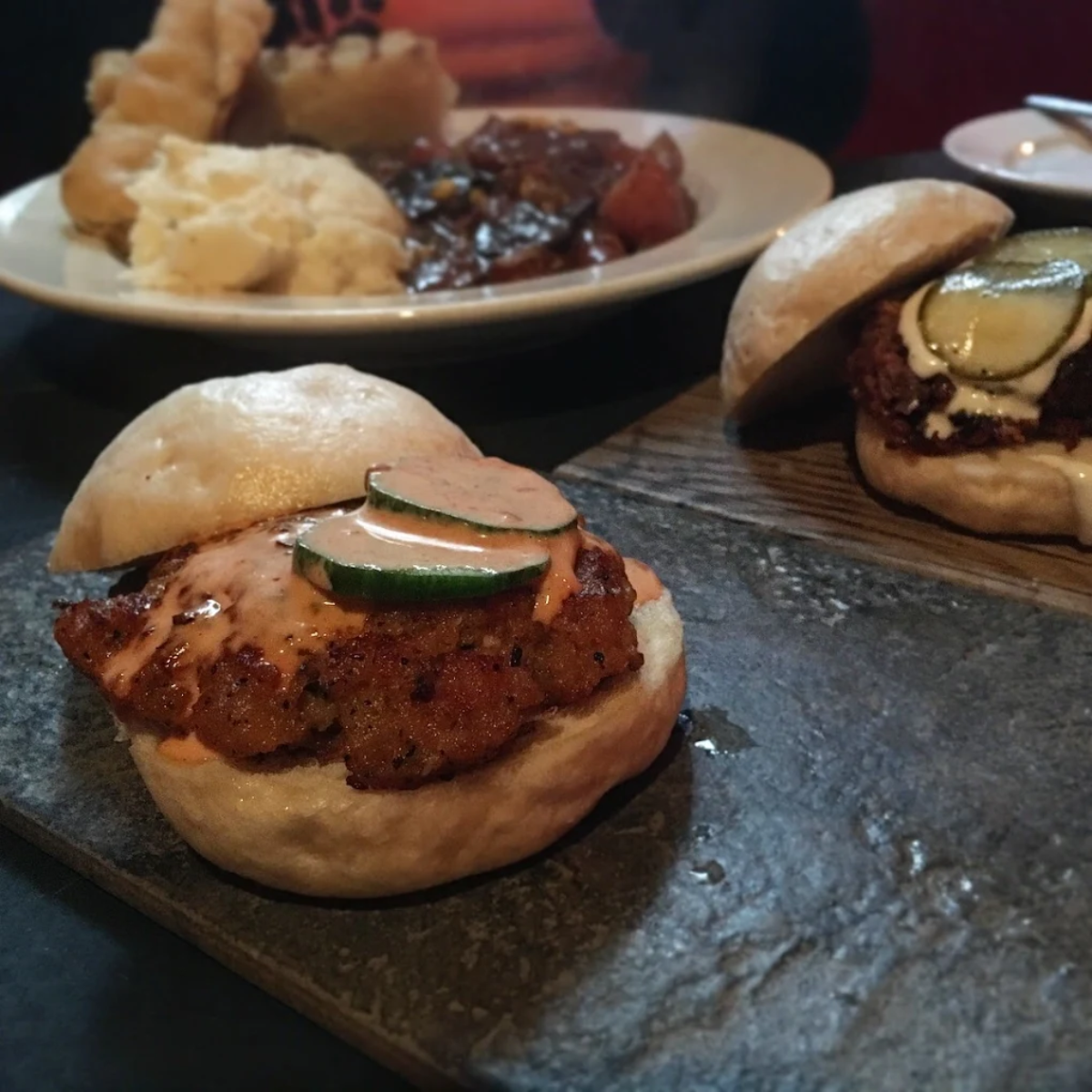 Close-up of two savory sandwiches with grilled patties, topped with cucumber slices and sauce, served on buns. A plate with mashed potatoes and a stew is blurred in the background.