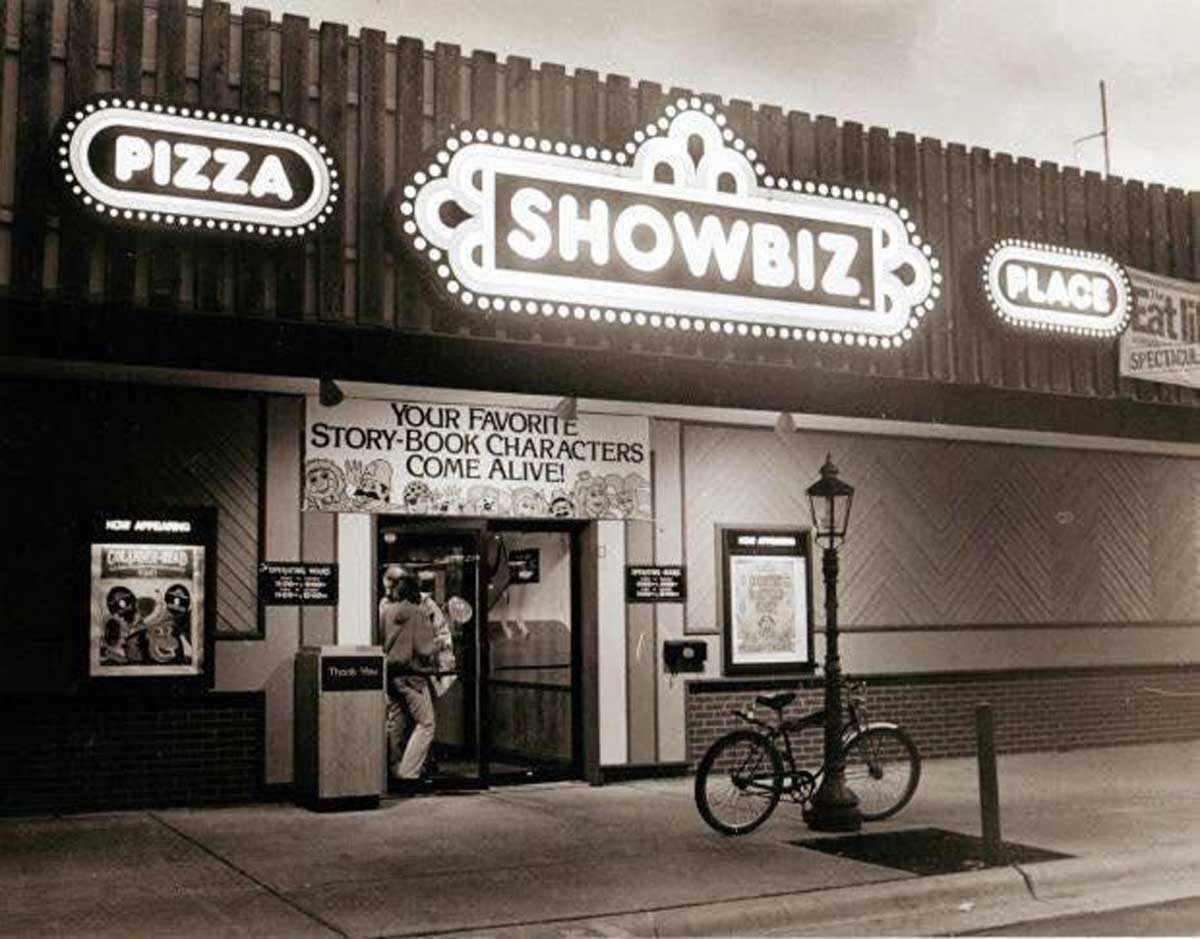 A vintage black-and-white photo of the exterior of a ShowBiz Pizza Place restaurant at night, featuring bright marquee signs and a bicycle parked outside near the entrance—a nostalgic look at one of America’s famous failed pizza chains.