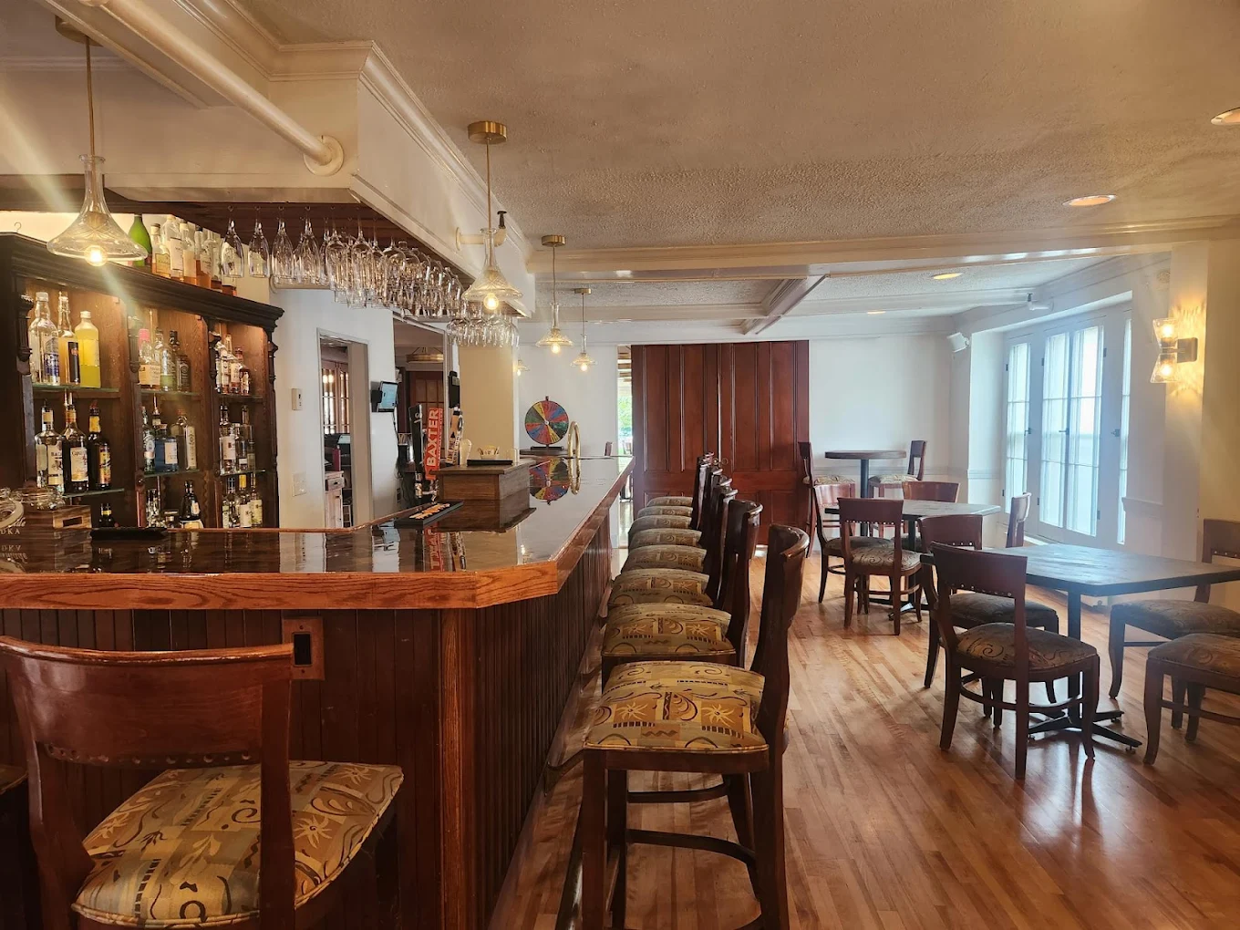 A cozy bar with a wooden counter, high bar stools, and shelves stocked with bottles. The room has wooden flooring, tables, and chairs, with bright lighting and large windows letting in natural light.