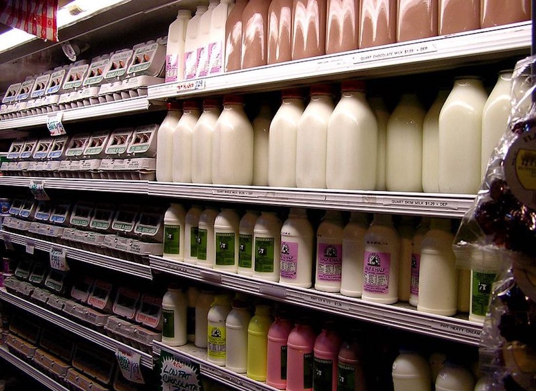 Refrigerated shelves stocked with cartons of eggs on the left and glass bottles of milk in various flavors, including white and chocolate, on the right. Some bottles have green and purple labels.