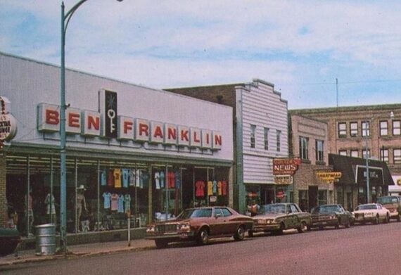 A vintage street scene shows the Ben Franklin store with large red letters, next to other small shops and parked classic cars lining the street under a blue sky.