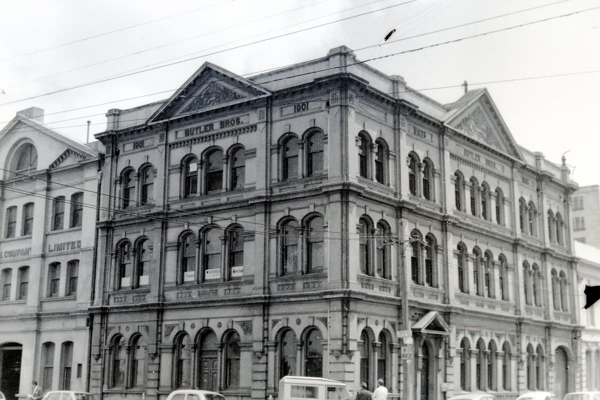 Black and white photo of a three-story historic building with arched windows and decorative stonework, once home to a five-and-dime store; signs read "Butler Bros. 1861." Several old cars are parked in front, with nearby buildings partially visible.