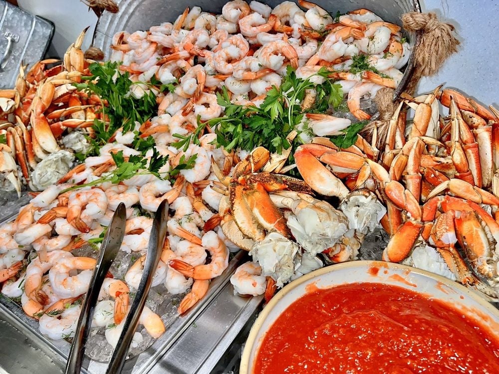 A seafood platter featuring piles of cooked shrimp and crab legs on ice, garnished with parsley, with metal tongs on the side and a bowl of red cocktail sauce in the foreground.