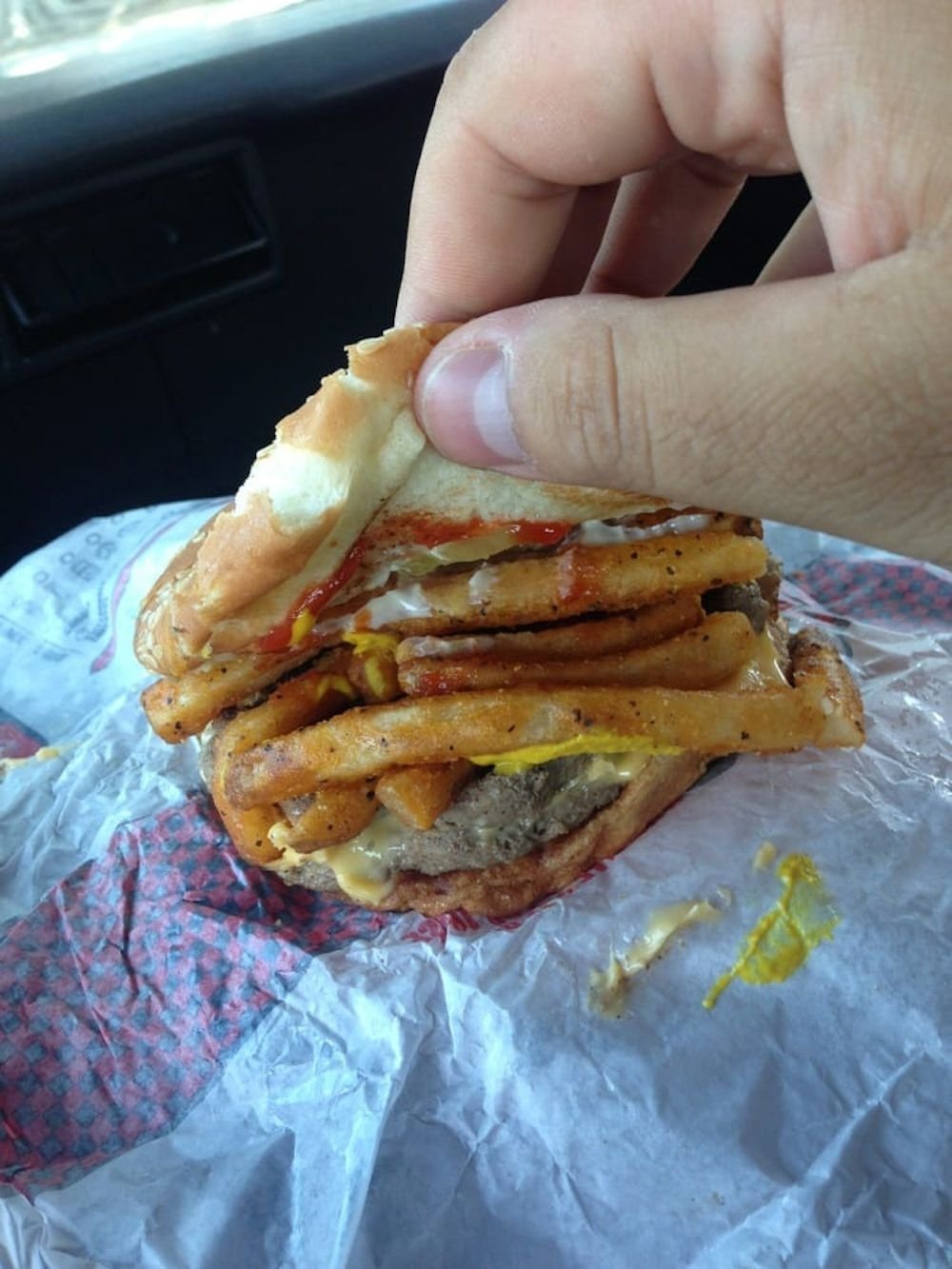 A hand holding a hamburger filled with French fries, ketchup, mustard, and mayonnaise, with a messy bun and wrapper underneath.
