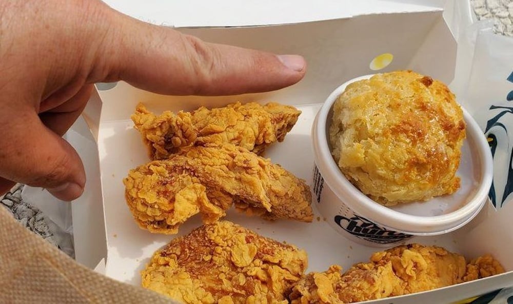 A hand points at a takeout box containing three pieces of fried chicken and a biscuit in a small cup. The food is crispy and golden-brown.