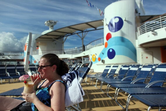 A woman in sunglasses relaxes on a lounge chair by a pool on a cruise ship, sipping a tropical drink with a small umbrella. Rows of empty blue chairs and colorful ship decor are visible under a sunny sky.