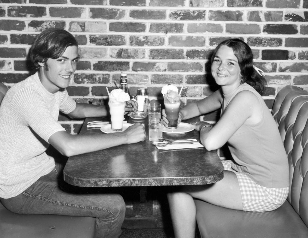 circa 1975: A teenage couple smiles as they sit in a booth at a diner, having ice cream and soda floats. There is a brick wall behind them.