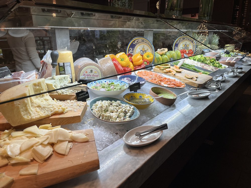 A buffet with various cheeses, cold cuts, salads, sliced bread, fresh vegetables, and colorful garnishes displayed under a glass cover. Small plates and serving utensils are placed along the counter.