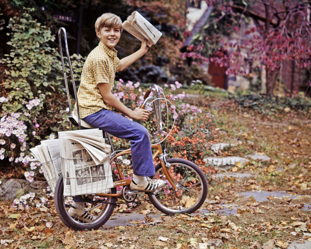 1970s newsboy on bike on suburban street with newspaper ready to toss for delivery.