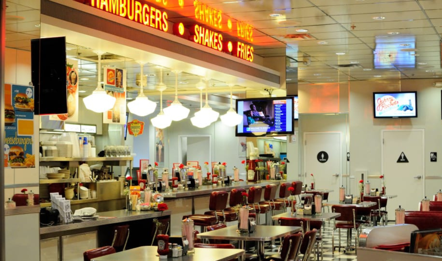 A retro-style diner with red chairs, small tables, a counter with stools, neon signs reading “HAMBURGERS,” “SHAKES,” and “FRIES,” and vintage décor on the walls.