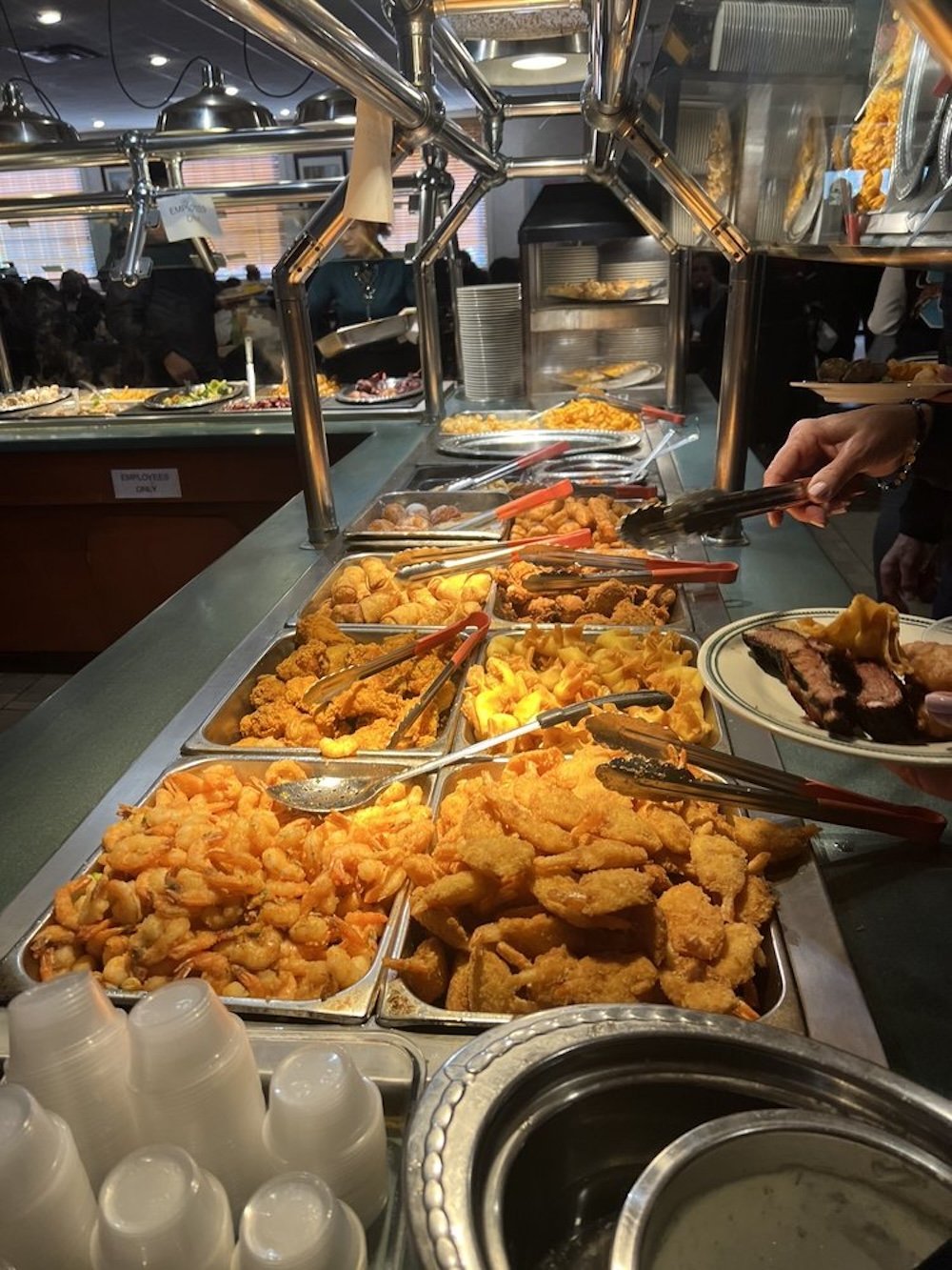 A buffet table filled with various fried foods, including shrimp, chicken, and other breaded items, with serving tongs and a person selecting food. Stacks of plastic cups and metal trays are visible in the foreground.