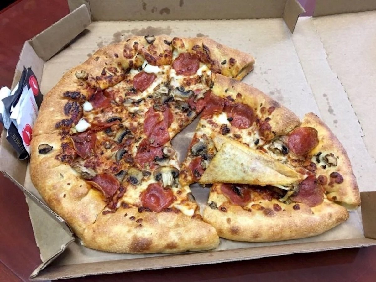 A partially eaten pizza with pepperoni, mushrooms, and melted cheese sits in an open cardboard box. One slice is flipped upside down, exposing its crust and sauce—a far cry from fare at the worst chicken chains. A dipping sauce container is in the corner of the box.