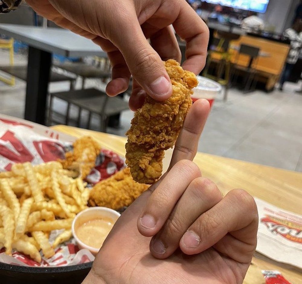 A close-up of two hands; one holds a fried chicken tender balanced on another person's upright finger. In the background, there are crinkle-cut fries and dipping sauce on a tray at a casual dining restaurant.