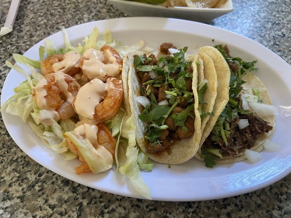 A plate with three tacos: one with shrimp, lettuce, and sauce; one with chopped meat, cilantro, and onions; and one with shredded beef, all on a white platter on a speckled table.