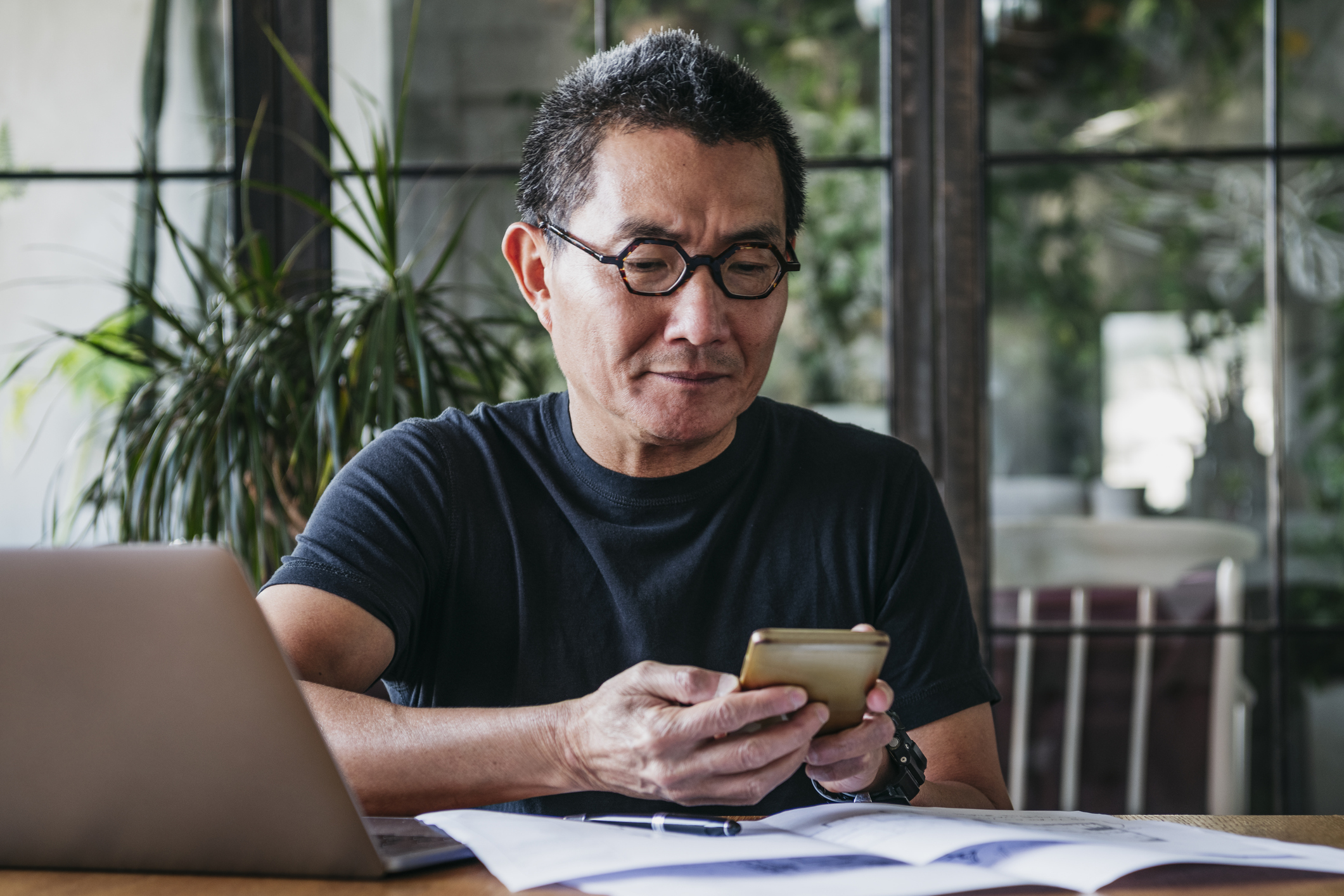Senior man texting on a smartphone in front of a laptop and paperwork at his desk in a modern living room with plants behind him and large window doors showcasing an enclosed garden area blurred in the background
