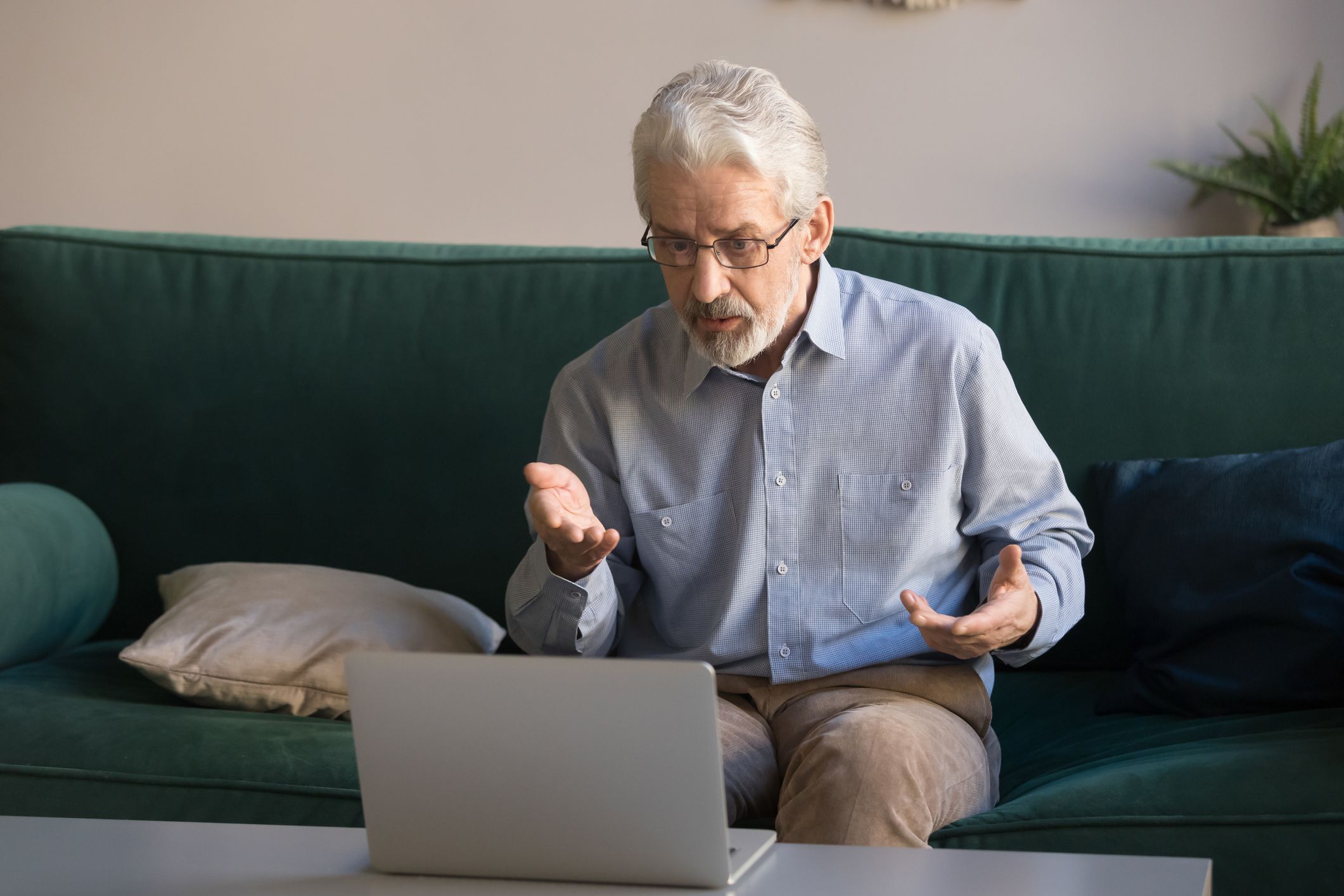 Senior man sitting on a sofa looking at laptop on adjacent coffee table, taking into it during a videocall with lavender colored wall in the background in living room