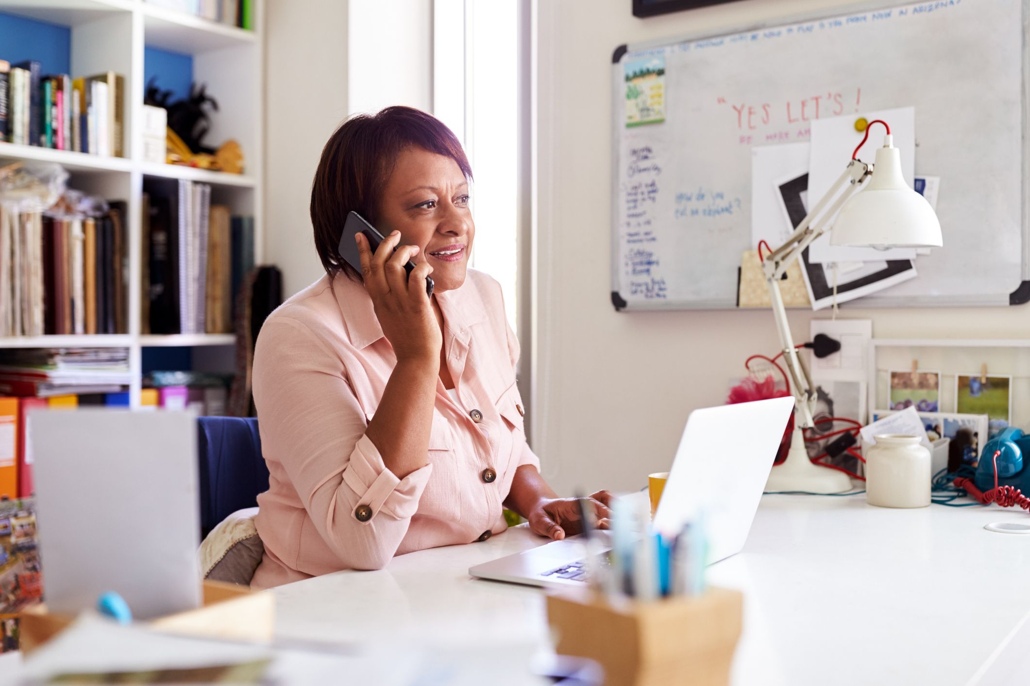 Senior woman on her smartphone, working with a laptop at a white desk in a trendy home office, with a whiteboard and several artsy items on her desk, with a cube bookcase in the background