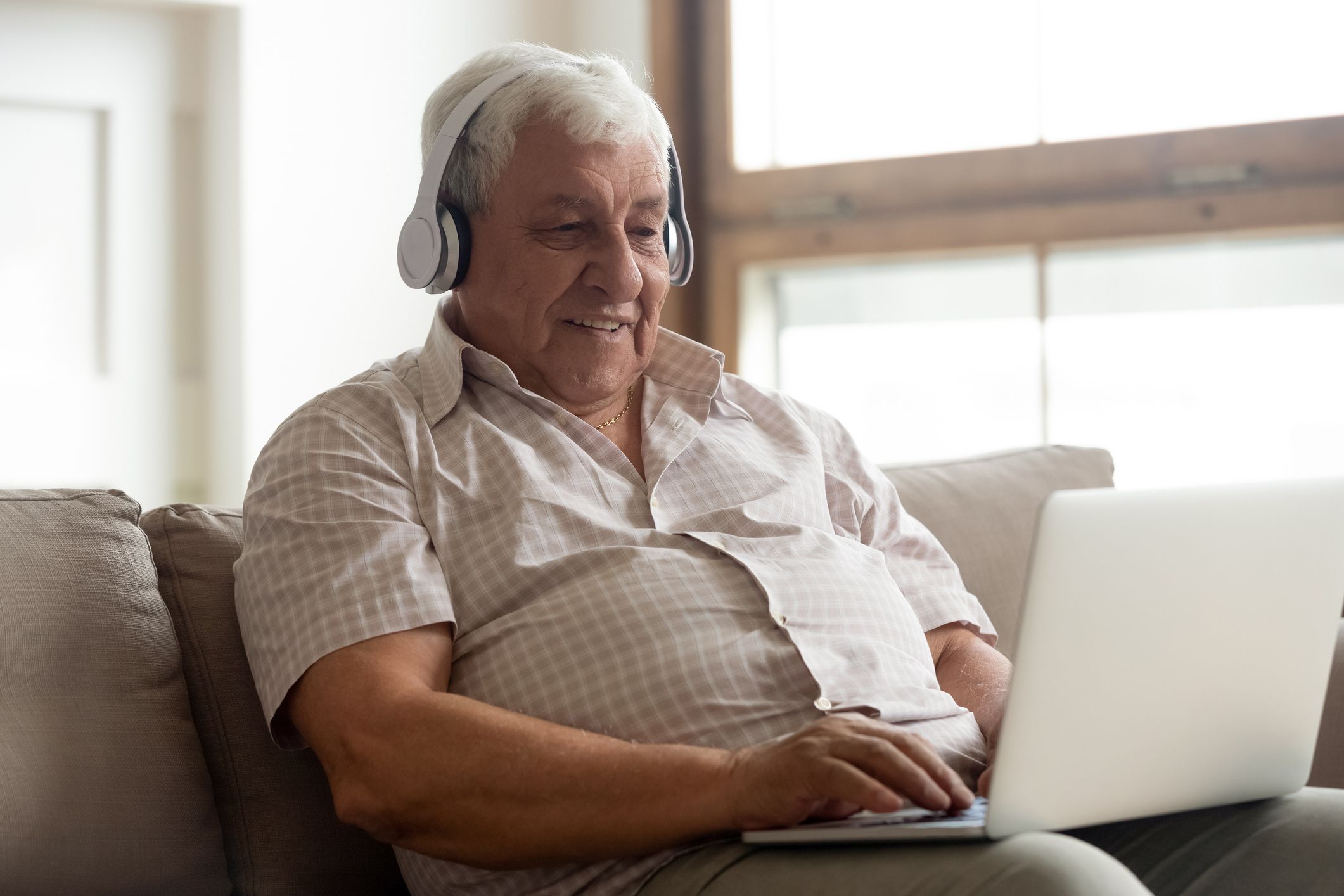 Smiling overweight senior man wearing headphones while on his laptop, sitting in a couch in his home, with a blurred background of a window and a white wall