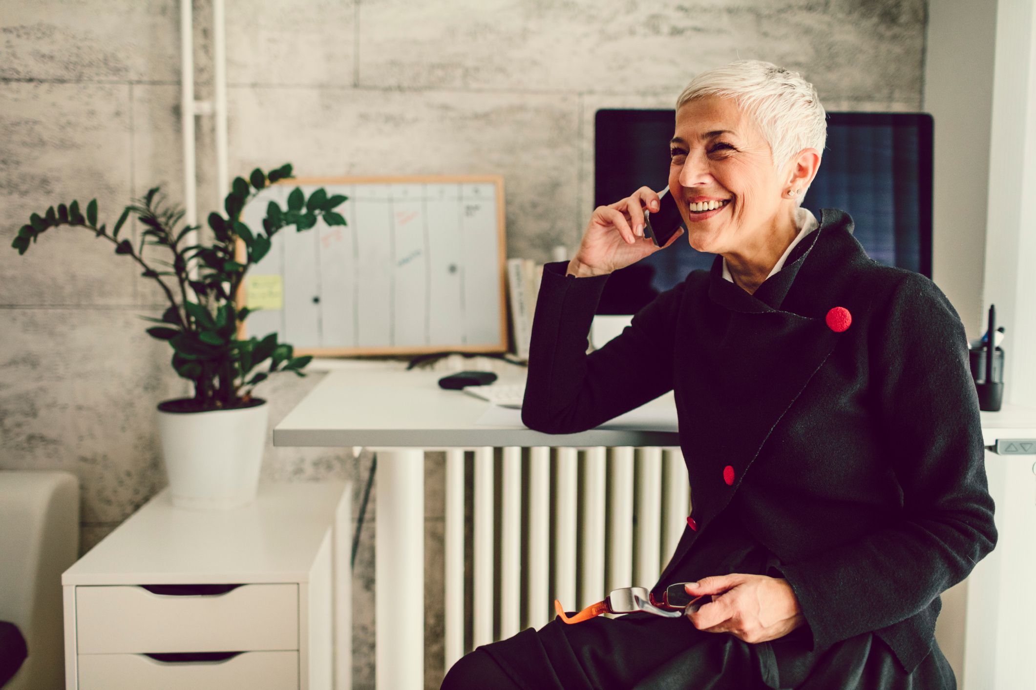 Smiling senior businesswoman talking on her smartphone while sitting away from a white desk with a desktop computer, a white board and such items, next to a matching white cabinet with a potted plant and a trendy grey wall