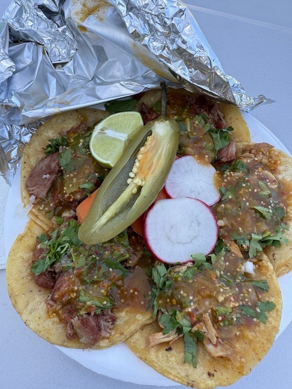 A plate with four tacos topped with meat, green salsa, chopped cilantro, and onions, garnished with sliced radishes, lime wedges, a jalapeño, and partially covered with aluminum foil.