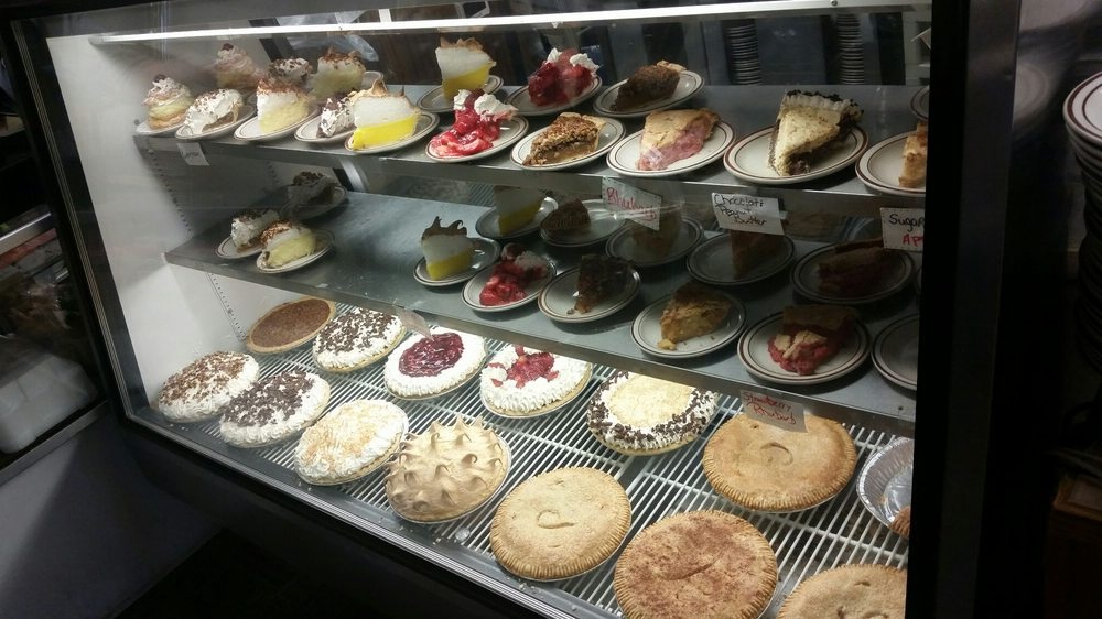 A glass display case filled with various whole pies on the bottom shelf and plates of individual pie slices, including fruit and cream varieties, on the upper shelves.