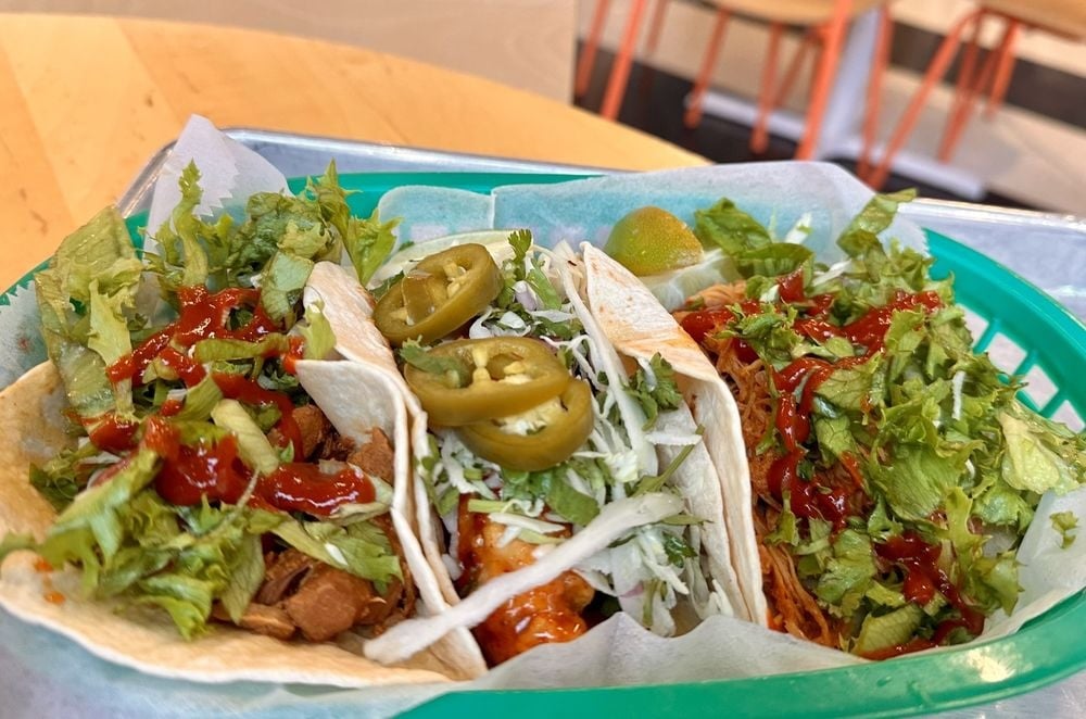 Three soft tacos in a green basket, filled with shredded lettuce, sliced jalapeños, diced meat, shredded cheese, and drizzled with red sauce. A lime wedge is placed on the side. Tables and chairs are blurred in the background.