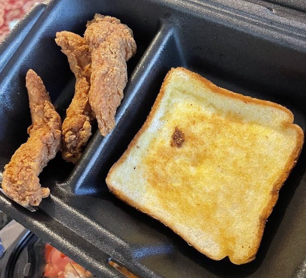 A black takeout container with three fried chicken tenders on the left and a single slice of toasted bread on the right.