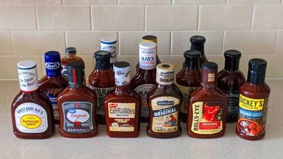 A variety of barbecue sauce bottles from different brands are arranged on a kitchen counter in front of a tiled backsplash.